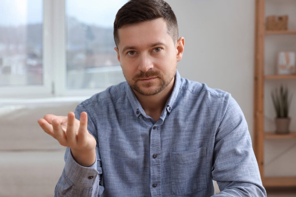 Man sitting on couch looking at the camera with a hand gesture.