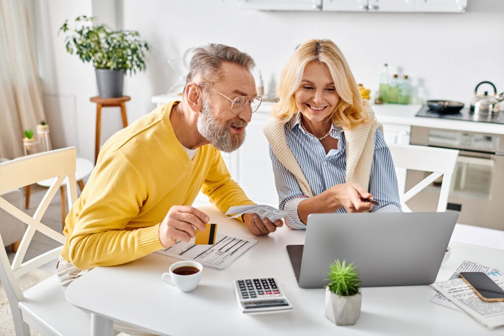 Couple in cozy homewear sit at a table, using a laptop and calculator.