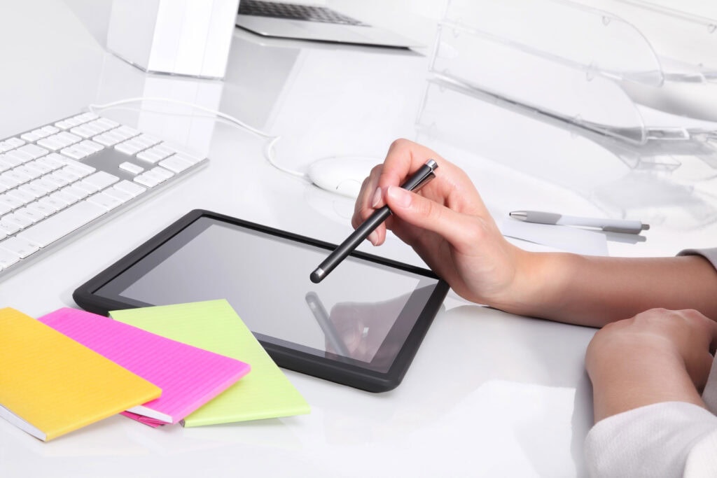 Person using a stylus on a tablet, surrounded by a keyboard, colorful notebooks, and another pen on a white desk.