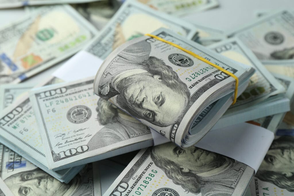 A bundle of U.S. hundred-dollar bills secured with a rubber band is placed on top of scattered bills.
