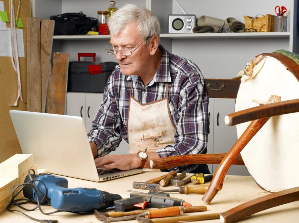 Senior craftsman sitting at his workshop using laptop. 