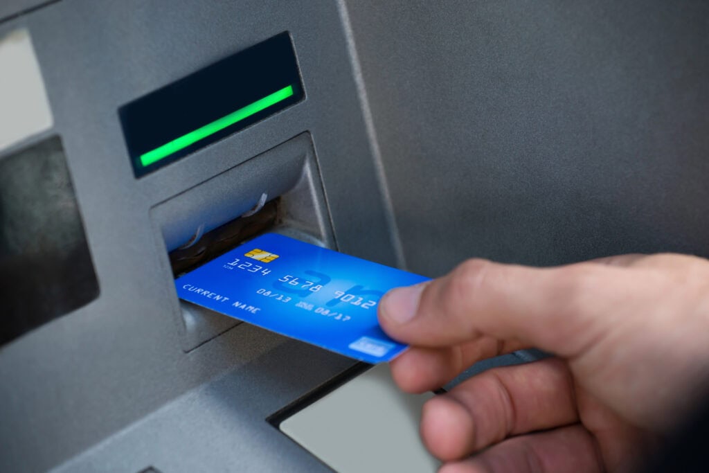 A hand inserts a blue credit card into an ATM slot displaying a green light.