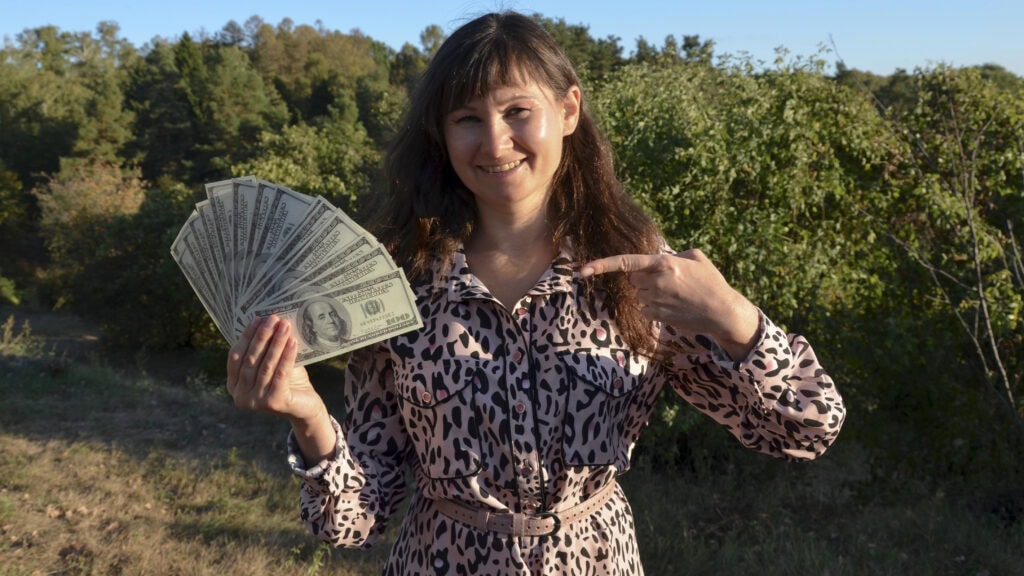 Woman points her forefinger to bunch of dollar, in outdoor setting. 