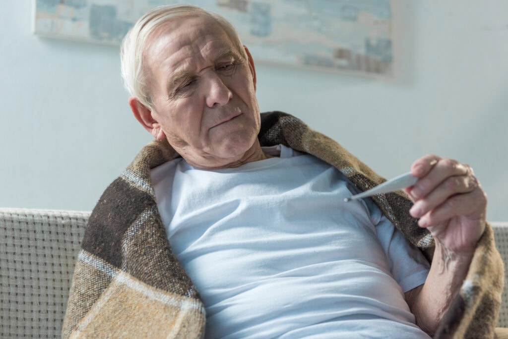 Senior sick man covered in plaid sitting on sofa and checking thermometer