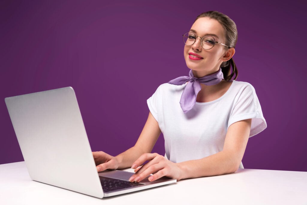Smiling girl using laptop and looking at camera isolated on purple