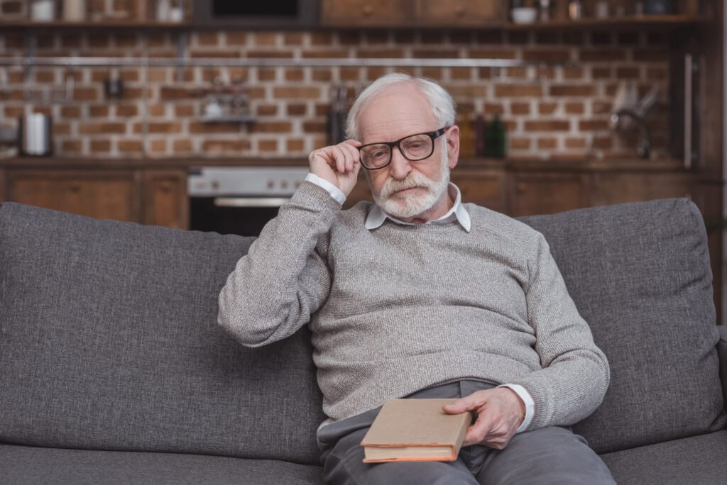 Handsome grey hair man touching glasses and holding book at home