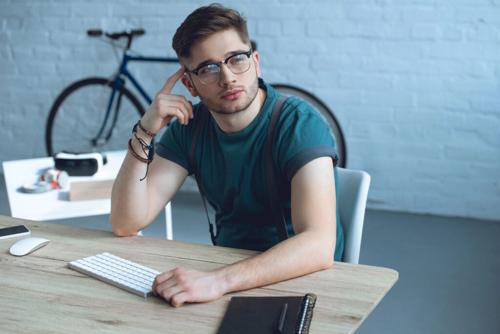 Handsome pensive freelancer in eyeglasses looking away while sitting at workplace