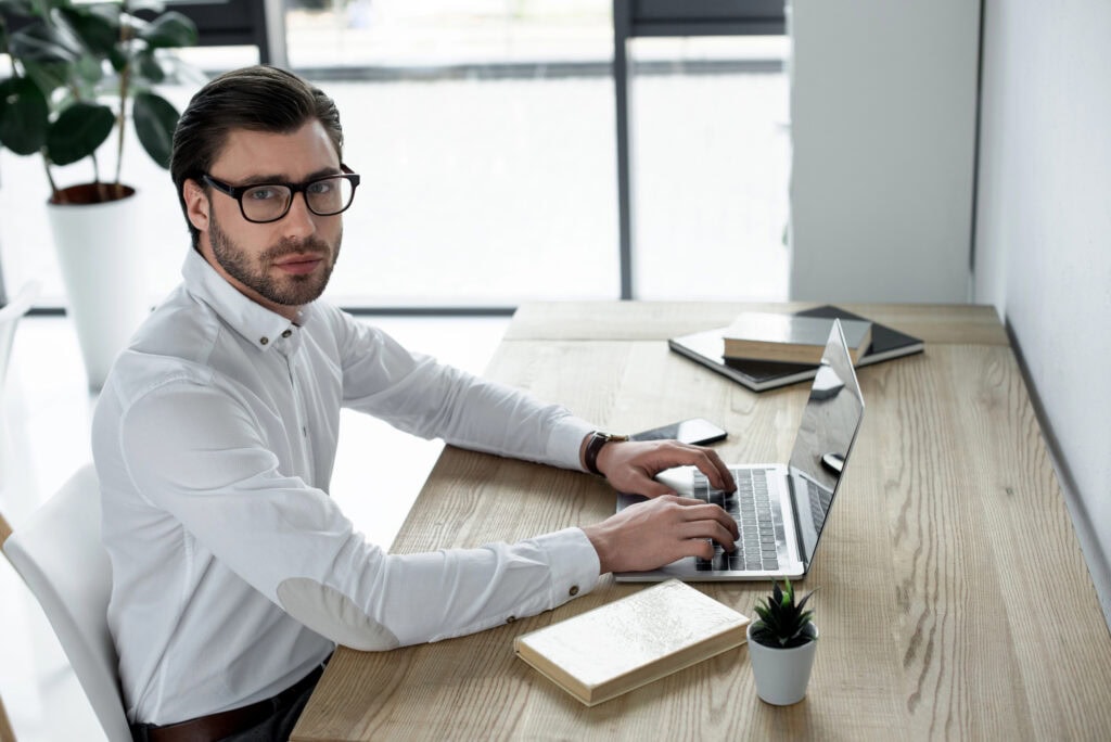 Confident young businessman working with laptop at modern office and looking at camera