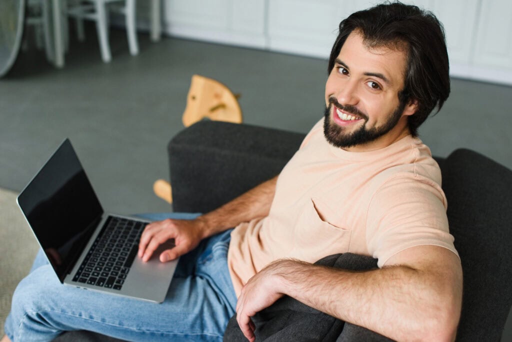 Side view of smiling man with laptop resting on sofa at home