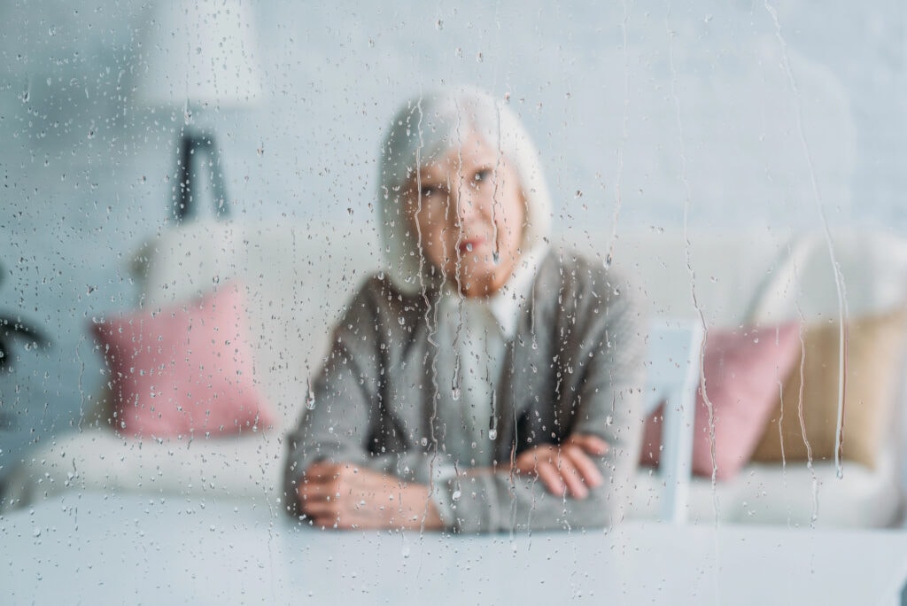 Selective focus of thoughtful senior woman in knitted jacket sitting at table alone at home