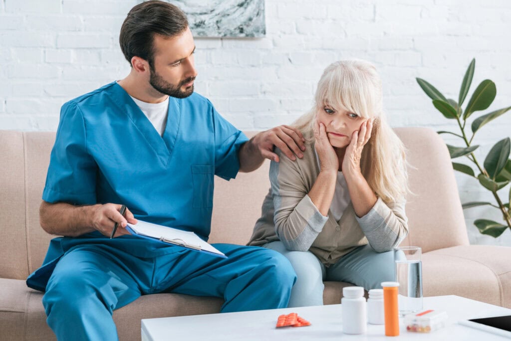 Male nurse holding clipboard and supporting upset senior woman looking at containers with pills