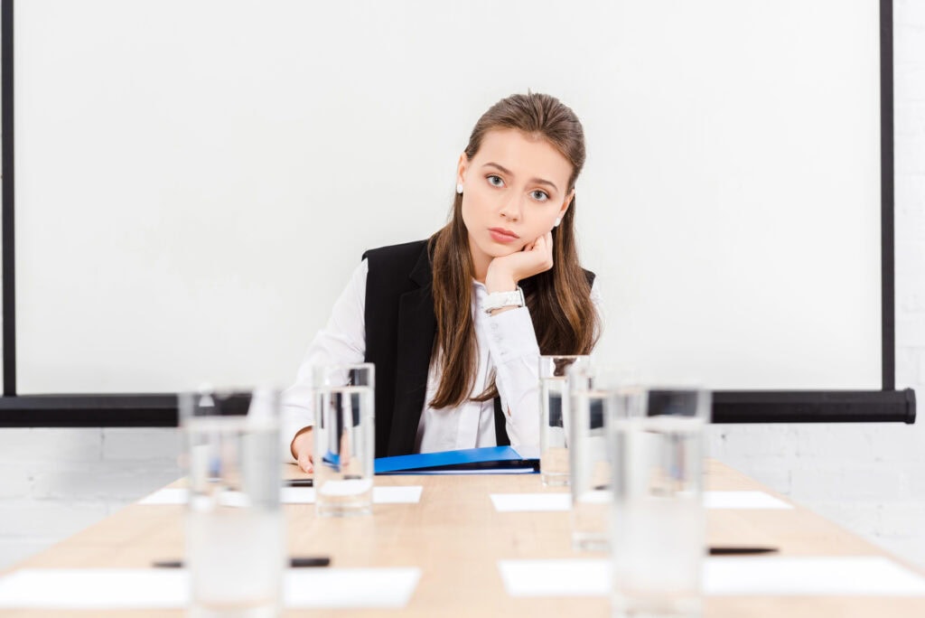 Bored young businesswoman sitting at table in conference hall at modern office and looking at camera