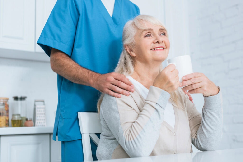 Cropped shot of caregiver standing near happy senior woman drinking tea in kitchen