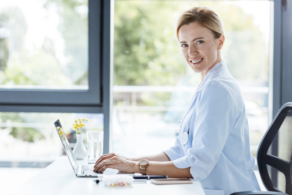 Smiling female doctor in white coat looking at camera and working on laptop at table in office