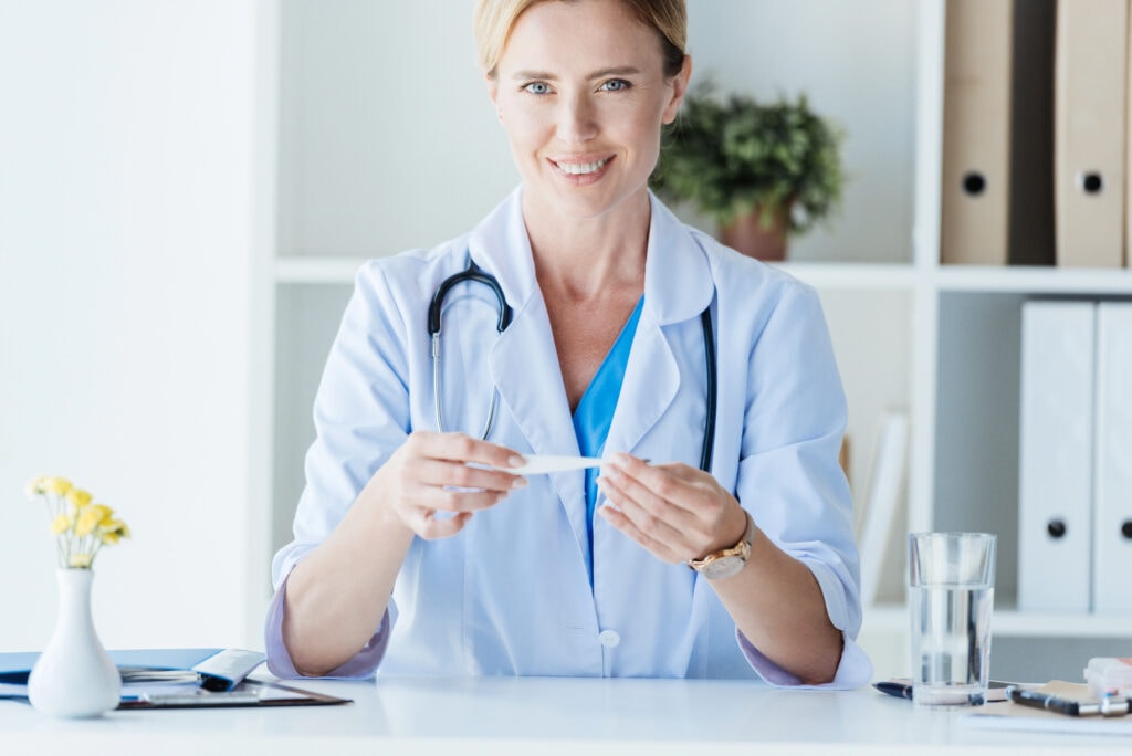 Attractive adult female doctor in white coat holding thermometer at table in office