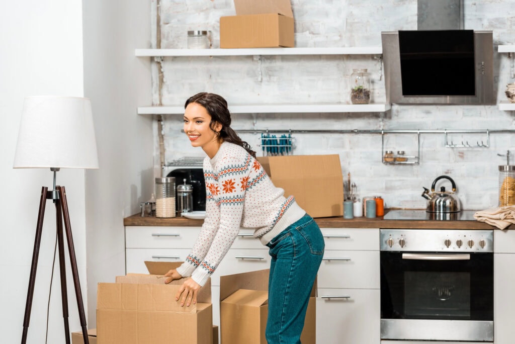 Attractive young woman standing near cardboard boxes in kitchen at new home
