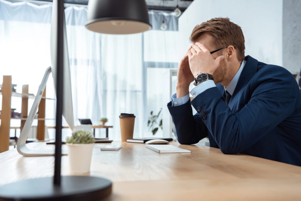 Upset businessman covering face by hands and working at table with computer monitor in modern office