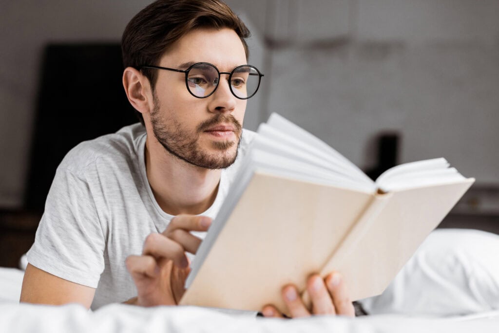 Handsome young man in eyeglasses lying in bed and reading book