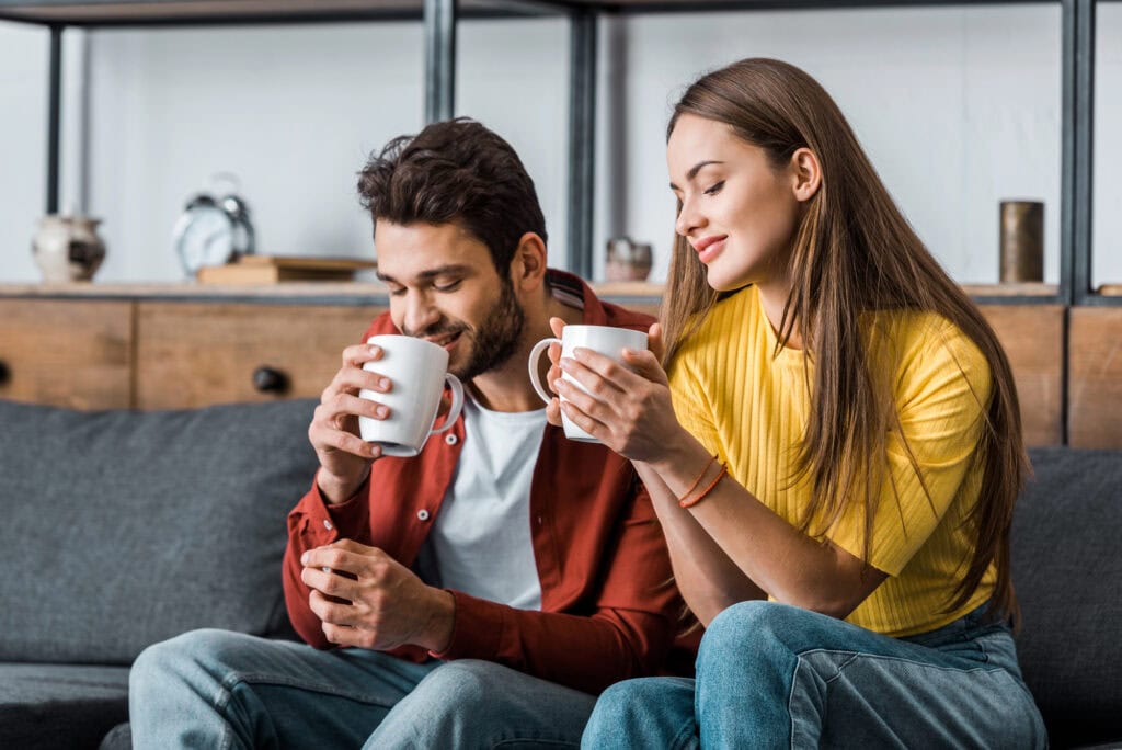 Cheerful girlfriend holding cup and sitting near boyfriend in living room