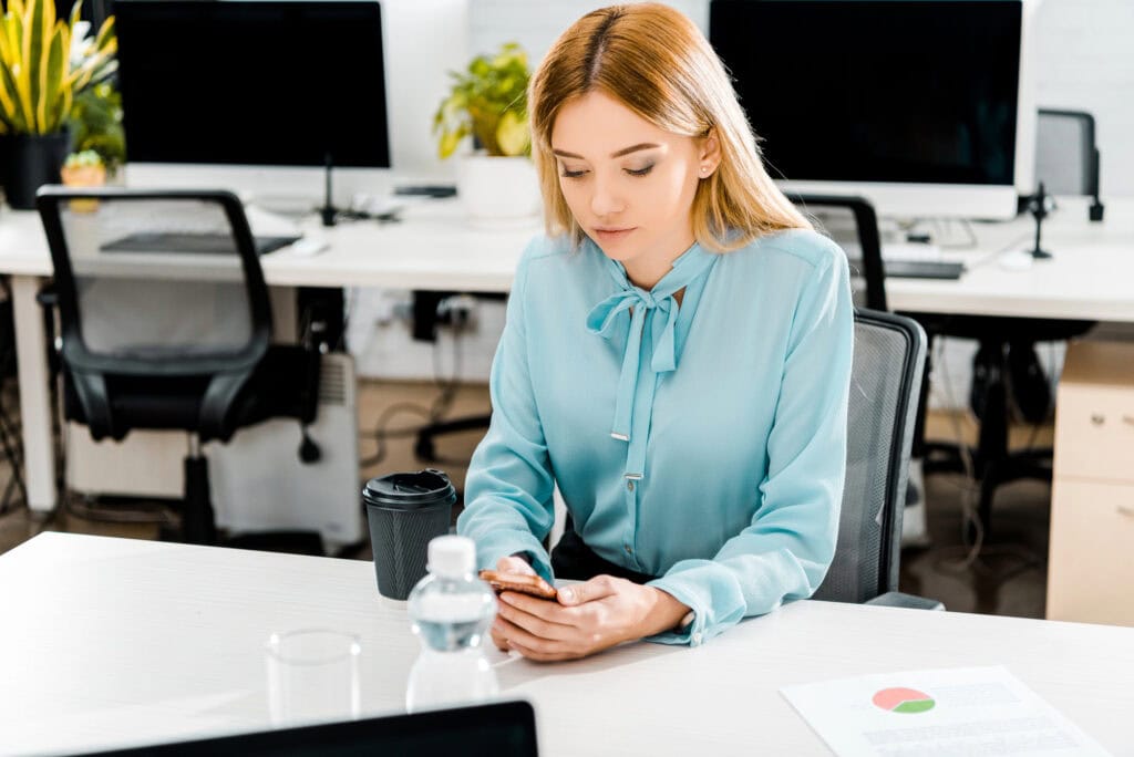 Portrait of young businesswoman using smartphone at workplace with coffee to go