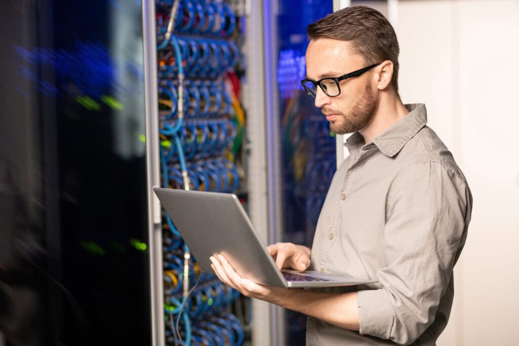 server engineer in glasses standing in database center and using laptop while troubleshooting problem