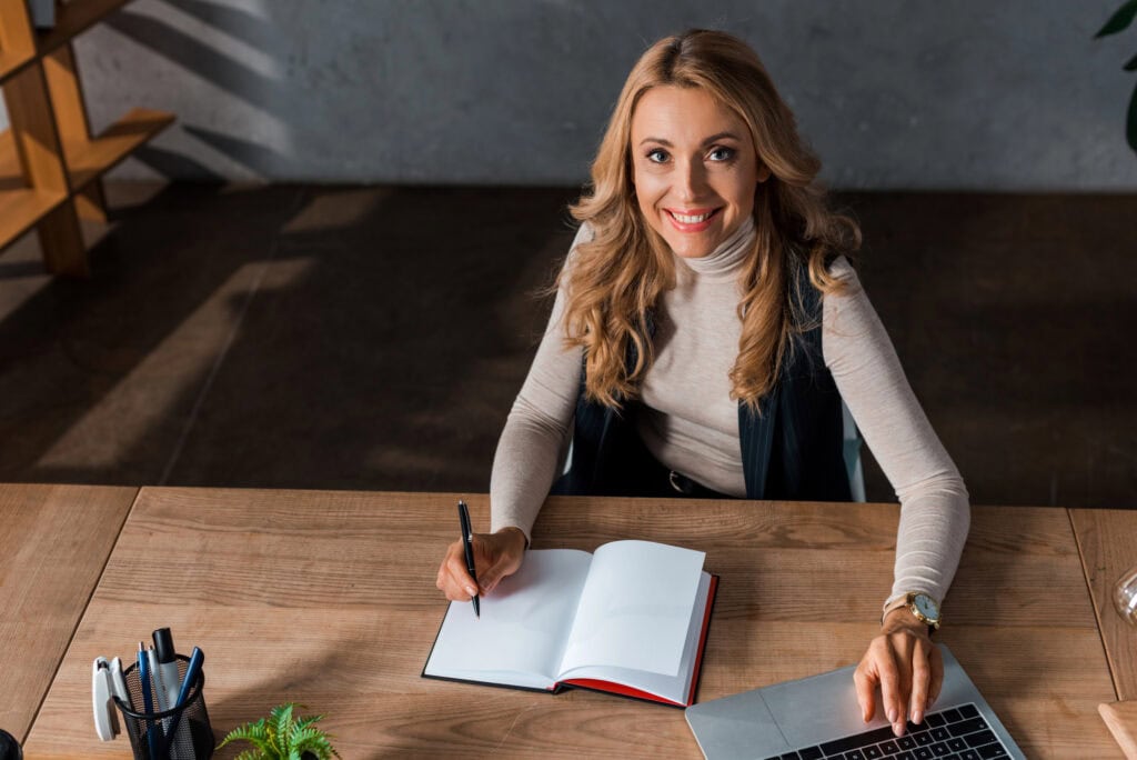 High angle view of attractive and blonde businesswoman smiling and sitting at table