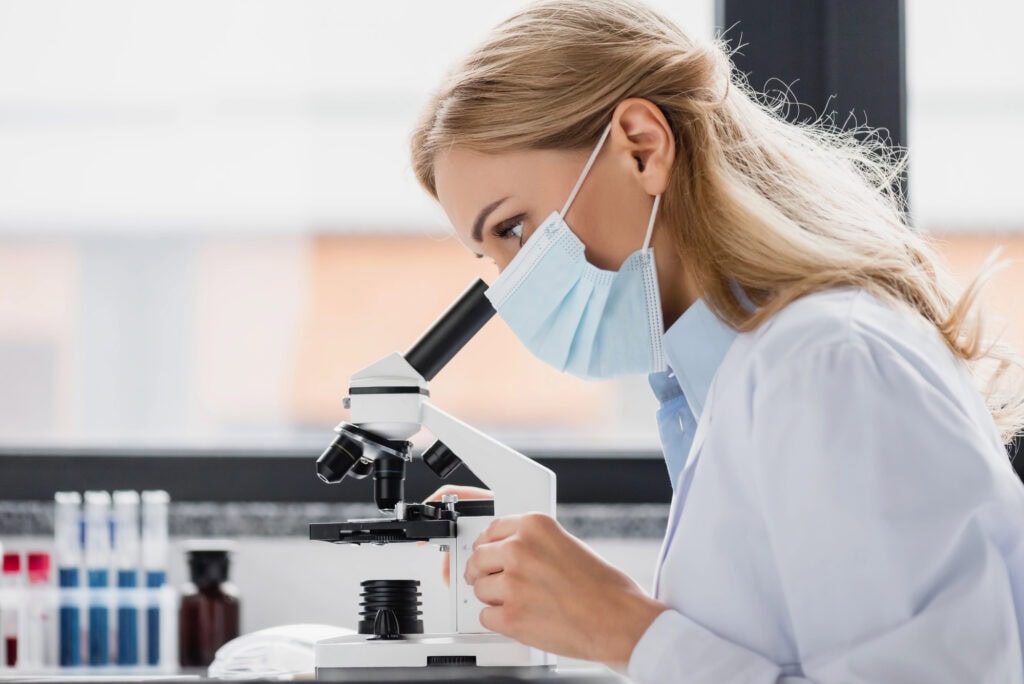 Specialist in medical mask looking through microscope in lab