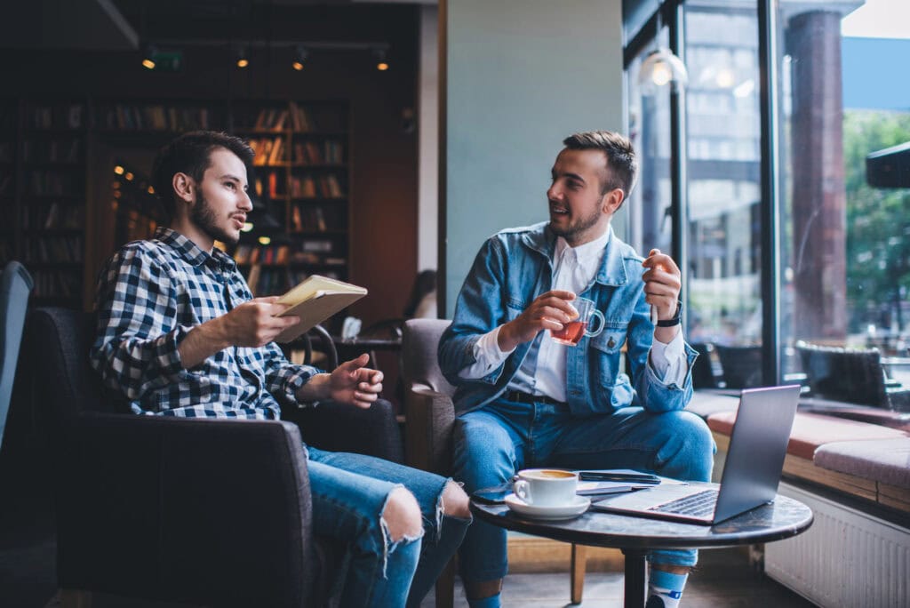 Cheerful smiling handsome men in casual outfit talking and drinking hot beverages while sitting in armchairs at table with laptop and notebook
