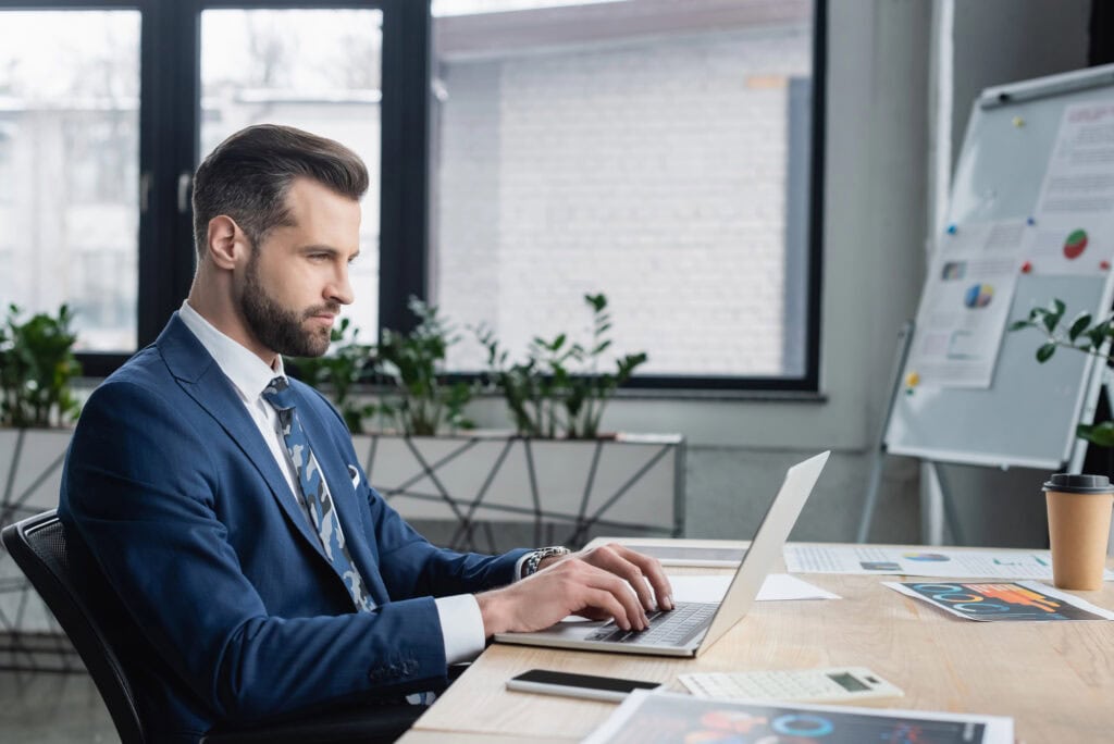 Economist typing on laptop near papers with analytics on work desk