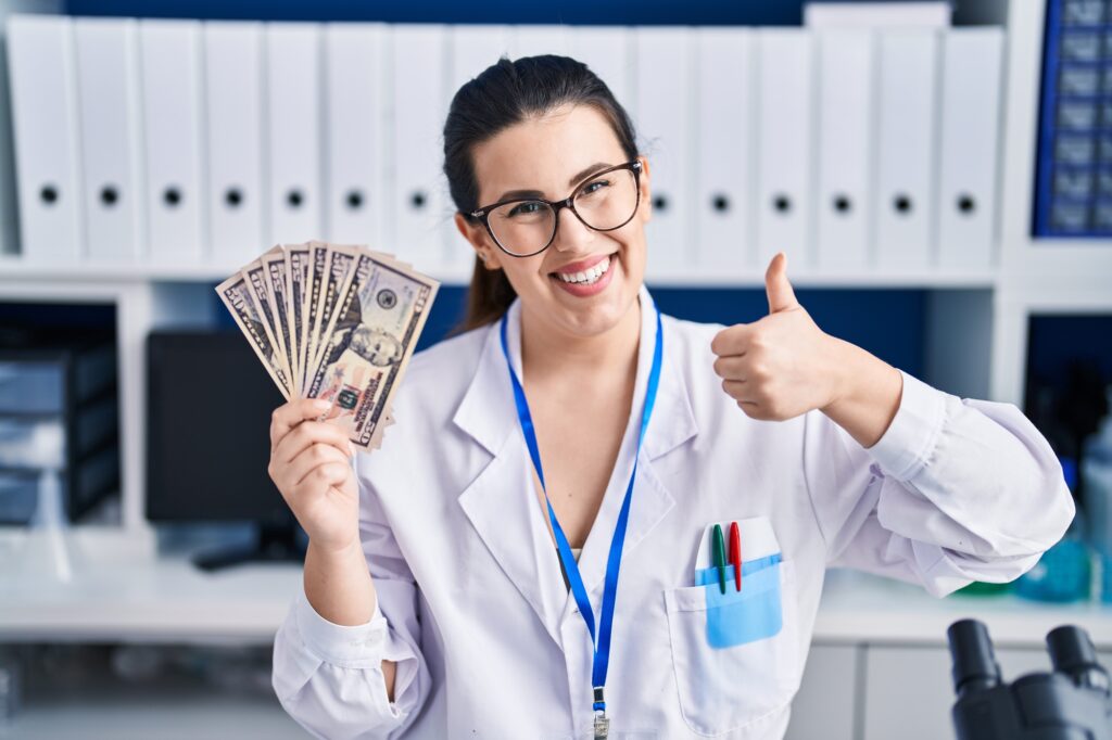 Woman working at scientist laboratory holding money dollar smiling happy, thumb up.