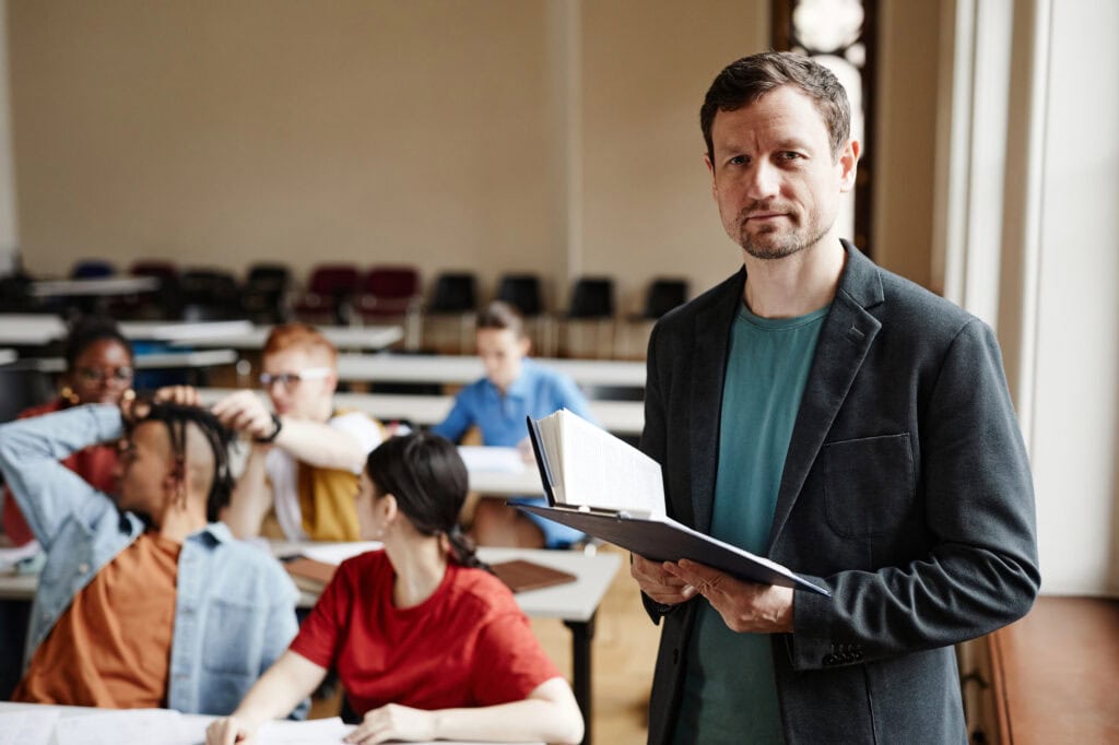 College professor looking at camera in classroom hall and holding book. 
