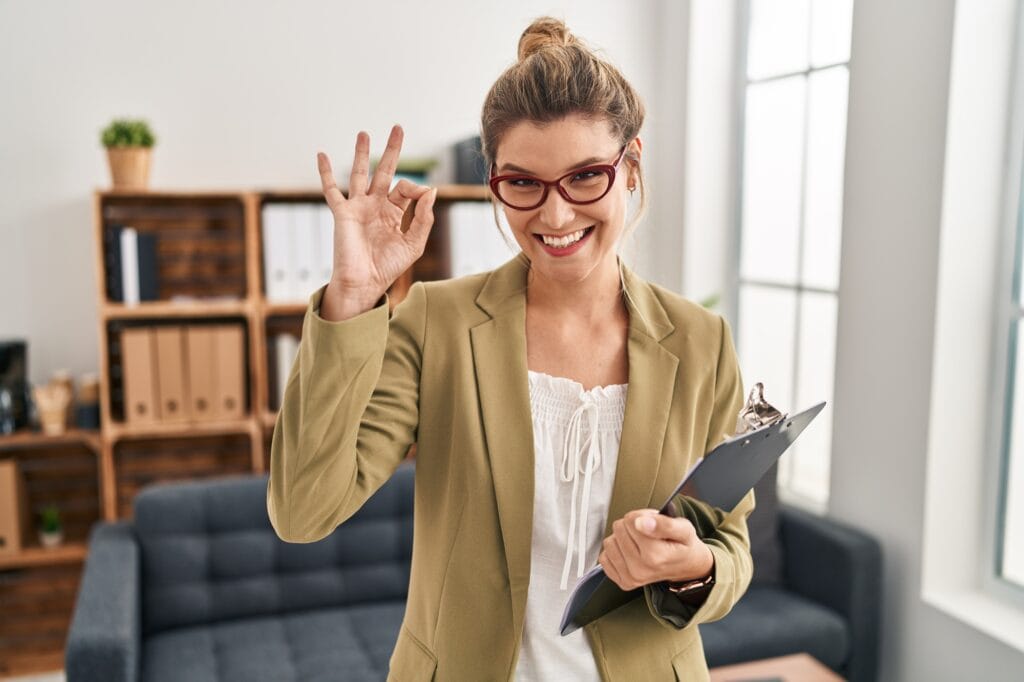 Woman working at consultation office smiling positive doing ok sign with hand and fingers. successful expression