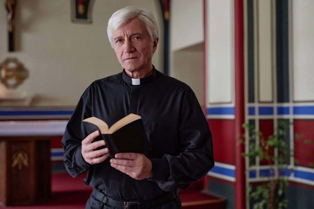 Senior priest looking at camera while standing with BIble book in church