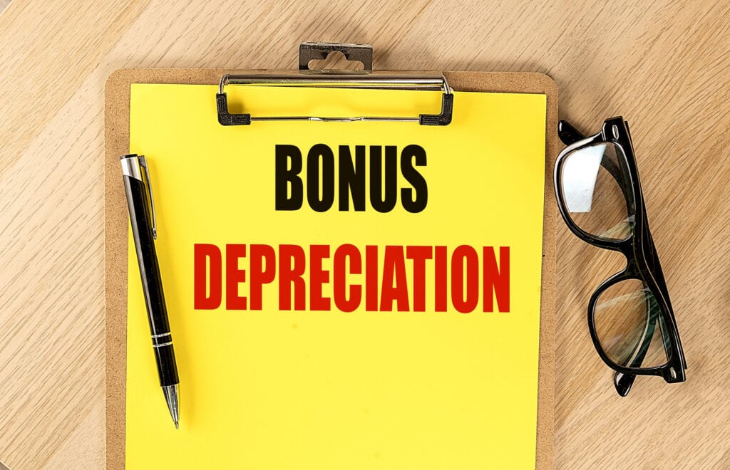 Clipboard with text bonus depreciation, pen, and glasses on a wooden table with a yellow background .