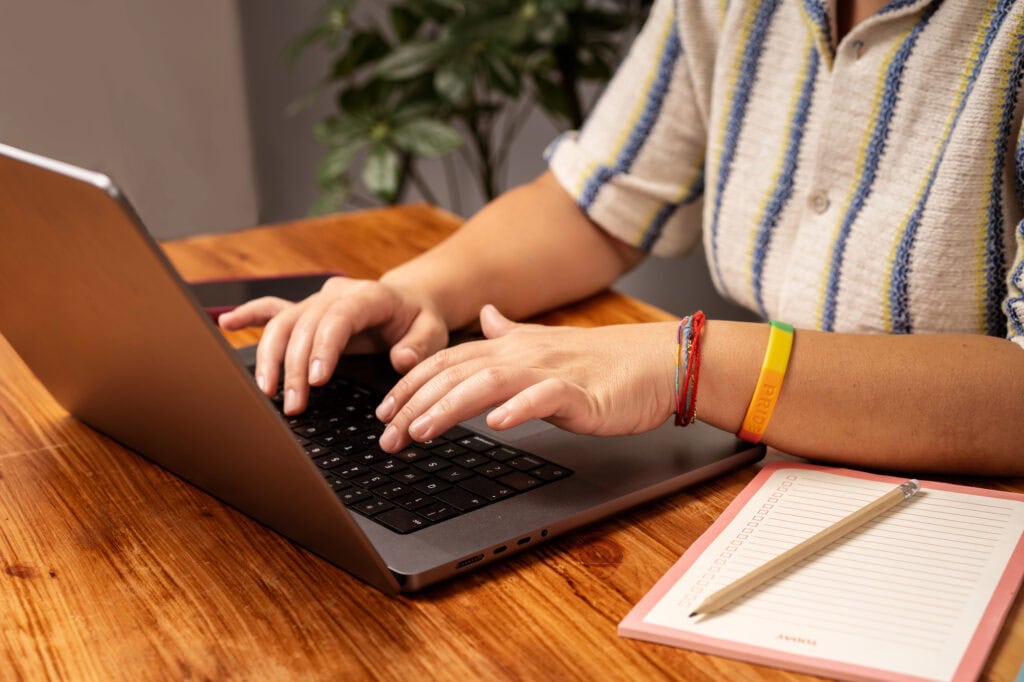 Close-up of hands typing on laptop at wooden desk with notepad