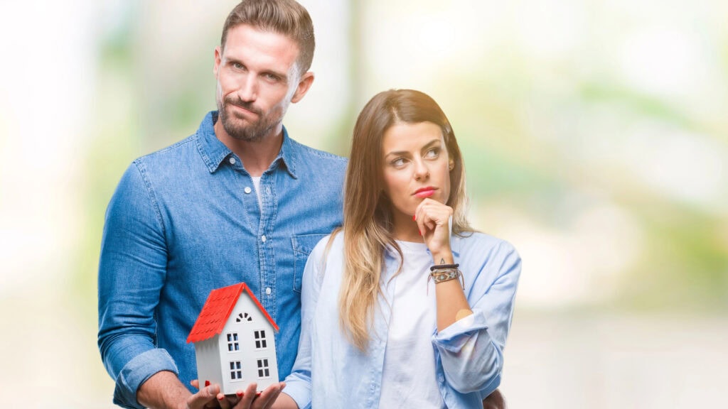 Young couple in love holding house over isolated background serious face thinking about question, very confused idea