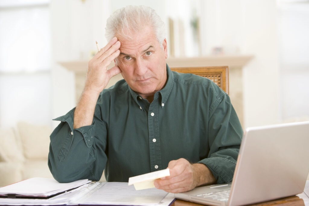 Man in dining room with laptop and paperwork looking frustrated